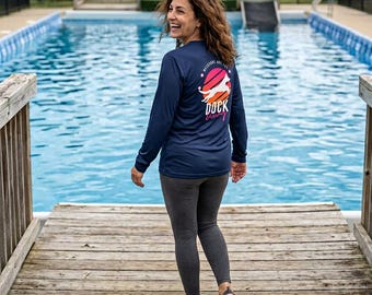 May include: Navy blue long-sleeve shirt with a graphic on the back that says "Weekends are for Dock Diving" with a dog silhouette. The person is wearing gray leggings and sandals, walking on a wooden dock near a pool.