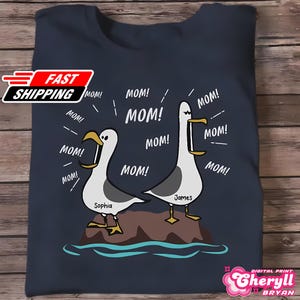 May include: Navy blue t-shirt featuring a cartoon illustration of two seagulls on a rock, each with a beak open and the word "MOM!" emanating from them. The seagulls are named Sophia and James. The shirt also has the text "FAST SHIPPING".