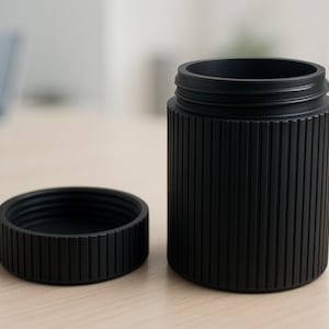 Puede incluir: Un recipiente cilíndrico negro con una tapa a juego. El recipiente tiene detalles verticales acanalados y una parte superior roscada. La tapa es circular con detalles acanalados similares. Los artículos están colocados sobre una superficie de madera de color claro.