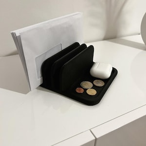 May include: A black desk tidy with three vertical dividers holding white envelopes. A small tray holds coins and a white wireless earbud case. The organiser sits on a white surface.