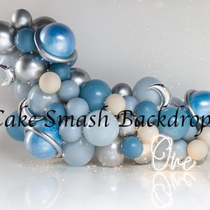 Puede incluir: Un arco de globos en tonos azules, plateados y crema, con detalles de planetas y lunas. Se ven las palabras "Cake Smash Backdrops" y la palabra "One" en neón. Adecuado para una fiesta de cumpleaños.