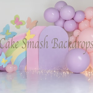 Könnte beinhalten: Pastellfarbener Hintergrund für die Fotografie mit Regenbogen, Luftballons und Schmetterlingsausschnitten. Der Hintergrund umfasst einen Regenbogenbogen, rosa und lila Luftballons und dekorative Schmetterlinge. Der Text "Cake Smash Backdrops" ist sichtbar.