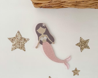May include: A pink felt mermaid with a purple crown and a white seashell bra, surrounded by gold glitter stars on a white background.