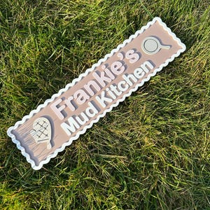 May include: A light brown wooden sign with white scalloped edges and white lettering. The sign reads "Frankie's Mud Kitchen". There are two white icons on the sign, one of a whisk and one of a frying pan.