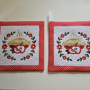 Peut inclure: Deux maniques carrées matelassées avec une bordure à pois rouges. Chacune présente un centre blanc avec un motif de tarte aux pommes brodé entouré d'une couronne florale. Une boucle en tissu blanc est attachée à chaque coin pour suspendre.