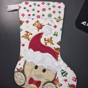 Könnte beinhalten: Ein weißer Weihnachtsstrumpf aus Stoff mit einem wiederkehrenden Muster aus Rentieren und Sternen. In der Mitte befindet sich ein großer, gestickter Lebkuchenmann mit einer roten Weihnachtsmütze mit weißem Kunstfellbesatz.