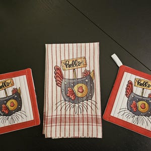 Op de afbeelding: Set van drie rode en witte keukendoeken met een geborduurd kipmotief in zwart en wit. De kip houdt een bord vast met de tekst "Hallo!"