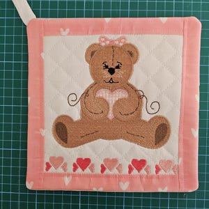 May include: Pink quilted pot holder with a brown teddy bear holding a heart. The teddy bear has a pink bow on its head and is surrounded by pink hearts.