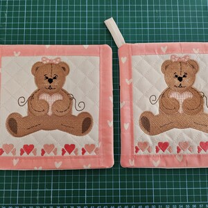 Peut inclure: Deux maniques matelassées rose et blanches avec un ours en peluche brun tenant un cœur rose au centre. L'ours en peluche a un nœud rose sur la tête et est entouré d'une bordure de cœurs rose et blancs.