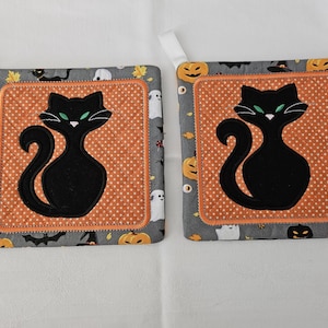 Könnte beinhalten: Zwei schwarze Katzen-Topflappen für Halloween mit orangefarbenen und weißen Punkten. Die Katzen haben grüne Augen und sitzen auf einem grauen Hintergrund mit Kürbissen, Fledermäusen und Geistern.
