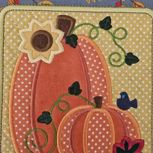 May include: A quilted fabric panel with an embroidered design of two pumpkins, a sunflower, and a small bird. The pumpkins are orange with white polka dots and green vines. The sunflower is yellow with a brown center. The bird is blue.