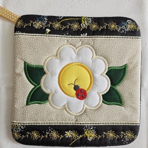 Può includere: Un sottopentola trapuntato beige e nero con un fiore bianco e una coccinella rossa ricamate. Il fiore ha un centro giallo e foglie verdi.