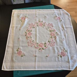 Op de afbeelding: Een vierkant wit stoffen tafelkleed met een centraal geborduurde krans van roze rozen en groene bladeren. De hoeken en randen zijn versierd met bijpassende rozenmotieven en een geometrisch randpatroon voor een symmetrisch ontwerp.