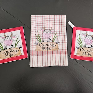 Könnte beinhalten: Zwei rote Topflappen mit einer Kuh und dem Text "Country Life" sowie ein rot-weiß kariertes Küchentuch mit einer Kuh und dem Text "Country Life", der darauf gestickt ist.