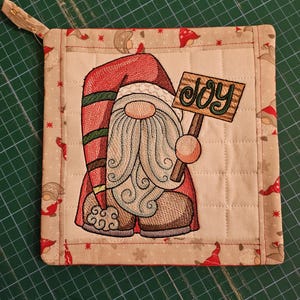 Op de afbeelding: Geborduurde kerstkabouter pannenlap met het woord "JOY" op een bord. De kabouter draagt een rode hoed en houdt een houten bord vast. De pannenlap is vierkant, met een lus om op te hangen.