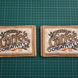 Puede incluir: Dos posavasos rectangulares de tela acolchada con bordes color canela y centro crema. Cada uno presenta un bordado en azul y marrón con el texto "POWERED BY Coffee AND CREATIVITY" rodeado de motivos decorativos de hojas y rodajas de naranja.