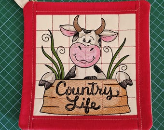 Op de afbeelding: Een vierkante stoffen pannenlap met een rode rand en een crèmekleurig middenpaneel met een geborduurde koe met een roze snuit. De koe staat achter een beige houten bordje met de zwarte geborduurde tekst Country Life.