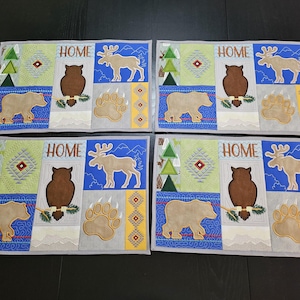 May include: Set of four gray placemats with a blue, green, and yellow geometric pattern. Each placemat features an embroidered design of an owl, a bear, a moose, and a paw print. The word "HOME" is embroidered on each placemat.