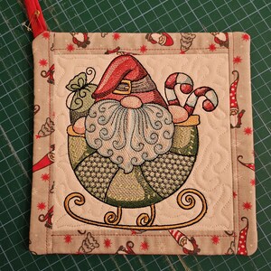 Op de afbeelding: Geborduurde kerstkabouter pannenlap met een rode lus om op te hangen. De kabouter draagt een rode hoed en heeft een lange witte baard, zittend in een slee met zuurstokken. De rand is beige met herhaalde kabouter- en sterpatronen.