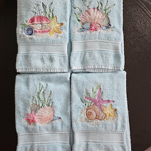 Puede incluir: Conjunto de cuatro toallas de mano azules con diseños bordados de conchas marinas, estrellas de mar y algas.