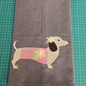 May include: A brown and tan dachshund wearing a pink and green dress with a flower on its collar. The dachshund is embroidered on a brown fabric kitchen towel.