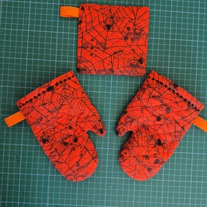 Könnte beinhalten: Ein Set aus zwei gesteppten Ofenhandschuhen und einem quadratischen Topflappen, alle aus leuchtend orangefarbenem Stoff mit schwarzem Spinnennetz- und Spinnenmuster. Jedes Teil verfügt über eine kleine orangefarbene Schlaufe zum Aufhängen.