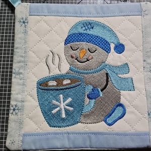 Può includere: Una presina trapuntata quadrata con un pupazzo di neve grigio ricamato che indossa un cappello e una sciarpa blu, che tiene in mano una tazza blu con un disegno di fiocco di neve bianco e vapore, su sfondo crema con bordi azzurri.