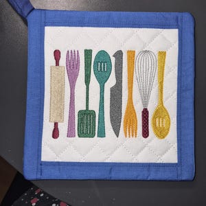 Puede incluir: Un agarrador de cocina cuadrado y acolchado con un borde azul y un panel central blanco. El centro presenta utensilios de cocina bordados en varios colores, incluyendo un rodillo, tenedor, espátula, cuchara, cuchillo, batidor y una cuchara de servir redonda.