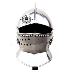 May include: A silver helmet with a visor and a crest on the top. The helmet has a decorative design around the visor and a small, pointed crest on top.