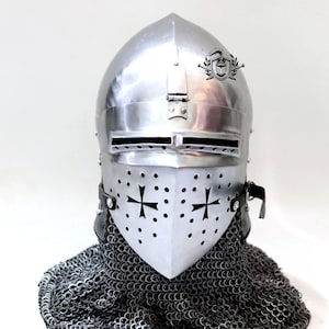 May include: A silver helmet with a pointed top and a face mask with a cross design. The helmet is worn over a chainmail coif.