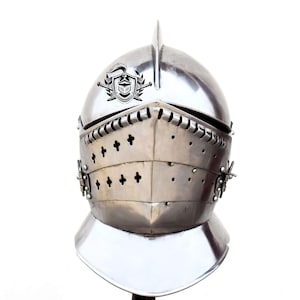 Puede incluir: Un casco de caballero plateado con una cresta y una visera. El casco tiene un diseño decorativo con pequeñas cruces en la parte delantera.