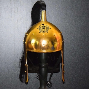 May include: A gold helmet with a black horsehair plume and a crest with a stylized design. The helmet has a pointed top and is decorated with small metal studs.
