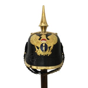 May include: A black leather Prussian Pickelhaube helmet with a gold eagle crest and a tall, pointed spike. The helmet has a brass trim and a leather chin strap.