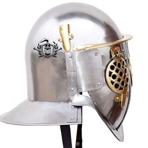 Pode incluir: Um capacete de prata com uma viseira de lat&atilde;o e uma crista decorativa no lado. O capacete tem uma m&aacute;scara facial de malha e uma correia de couro.