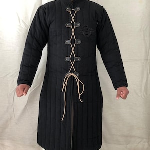 May include: A black quilted jacket with a long, fitted style. It has a high collar and a lace-up front with leather straps and buckles. The jacket is lined with a cream-colored fabric.