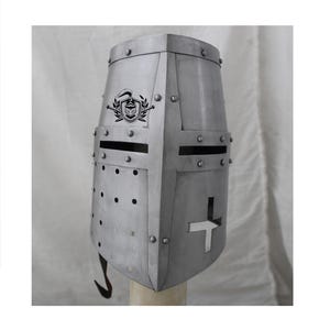 Op de afbeelding: Een zilveren helm met een kruisvormige uitsparing aan de voorkant en een schild met een zwaard en een helmdesign aan de bovenkant. De helm heeft klinknagels en een kleine haak aan de onderkant.