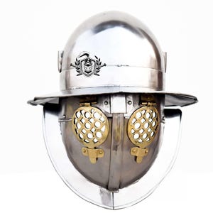 Puede incluir: Un casco de plata con una visera de latón y un emblema decorativo en la parte superior. El casco tiene un ala ancha y una visera con bisagras que se puede subir o bajar.