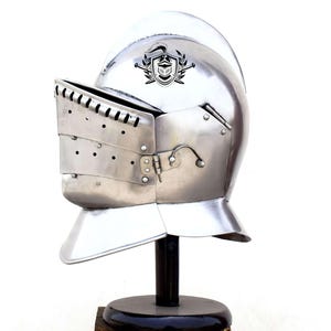 May include: A silver knight's helmet with a visor and a crest on the top. The helmet is on a black stand.