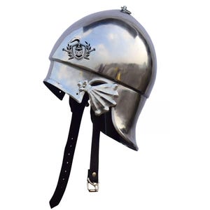 May include: A silver helmet with a crest and a leather strap. The helmet has a pointed top and a curved brim. The crest is a stylized eagle with wings spread. The strap is attached to the back of the helmet and has a buckle.