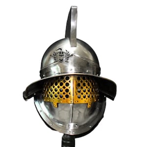 Könnte beinhalten: Ein silberner Gladiatorenhelm mit einem goldfarbenen Drahtgitter-Gesichtsschutz und einer Aufschrift auf der Oberseite.