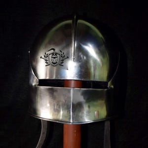 May include: A silver helmet with a black and white emblem of a knight with a sword and shield. The helmet has a visor and a neck guard.