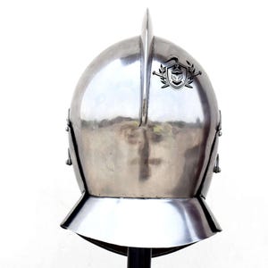 May include: A silver helmet with a pointed top and a decorative crest on the front. The helmet has a visor that can be raised or lowered.