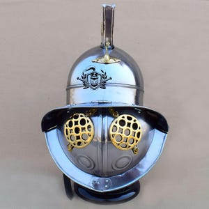 May include: A silver gladiator helmet with a gold crest and eye guards. The helmet has a tall, pointed crest and a face mask with two round, open eye guards.