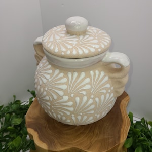May include: A beige ceramic sugar bowl with a lid, adorned with white floral patterns. The bowl has two handles and sits on a wooden pedestal. The lid features a small, round knob.
