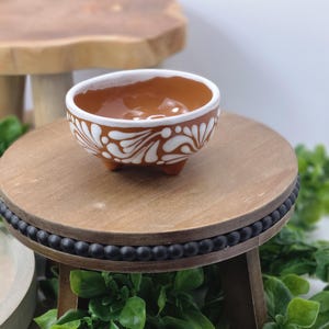 May include: A small, brown ceramic bowl with white floral designs and a white rim. The bowl sits on a wooden stand with black beaded trim. The background includes a wooden table and greenery.