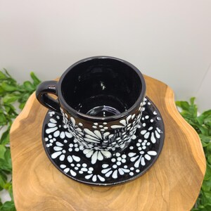 May include: Black ceramic teacup and saucer with a white floral design. The cup has a handle and rests on a matching saucer. The set is presented on a wooden surface, giving a rustic or handmade feel.