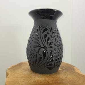 May include: A dark gray ceramic vase with a glossy black rim and embossed floral design. The vase is displayed on a natural wood surface, showcasing its decorative details and contrasting textures. A stylish home decor item.