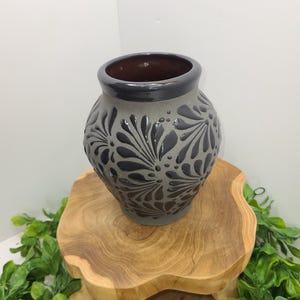 May include: A gray ceramic vase with a dark rim and interior, featuring a raised floral pattern. The vase is displayed on a wooden pedestal with a natural wood grain finish, surrounded by green foliage.