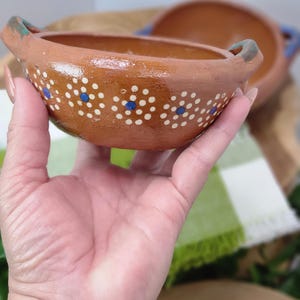May include: A handmade, terracotta-colored bowl with two handles. The bowl is decorated with white and blue polka dots. Another bowl is visible in the background. The bowl is held by a person.