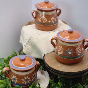 May include: Three terracotta-colored ceramic pots with lids. Each pot has a round body, two small handles, and a lid with a blue rim and knob. The pots are decorated with white and blue floral and geometric patterns.